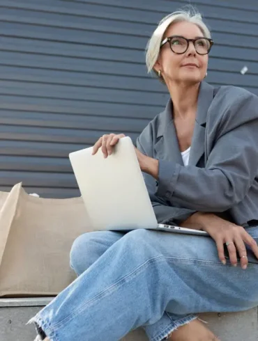 Oudere dame met jeans zit buiten met tas en laptop
