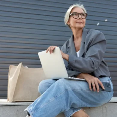 Oudere dame met jeans zit buiten met tas en laptop
