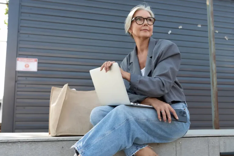 Oudere dame met jeans zit buiten met tas en laptop